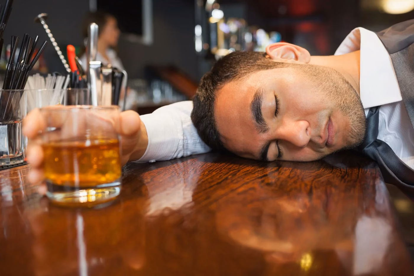A man asleep on a bar counter beside a glass of whiskey